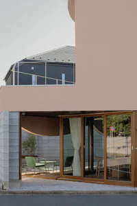 Modern residential building with a soft pink facade, large wooden-framed glass sliding doors, and an outdoor patio furnished with green metal chairs and a small table, next to a dark brick building in the background.