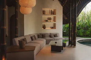 A contemporary open-air living room in a Bali villa featuring a large L-shaped sectional sofa, glass coffee table, built-in illuminated niches with pottery and artifacts, hanging paper lanterns, and dark bamboo structural columns overlooking a curved swimming pool and lush tropical garden.