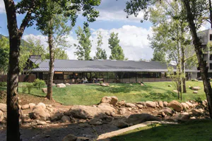 Modern single-story restaurant building with dark corten steel roof and full-height glass walls, situated beside a rocky stream and surrounded by lush trees and lawn.