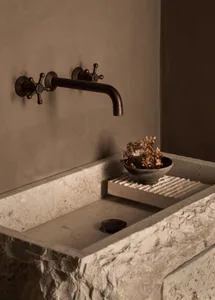 Close-up of a rustic travertine utility sink with a dark bronze wall-mounted faucet featuring cross handles, a small ceramic bowl with dried hydrangeas resting on a ribbed stone ledge, set against a textured plaster wall.