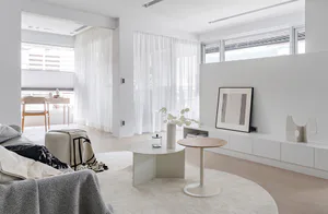 Modern minimalist living room with white curved nesting coffee tables, round white rug, low white media console, framed abstract art, sheer white curtains, and a grey textured sofa in the foreground.