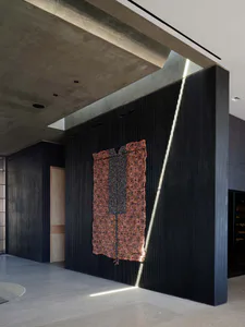 Modern minimalist concrete hallway featuring a large black fluted wall with dramatic linear LED lighting and a vibrant terracotta and black bead tapestry artwork.