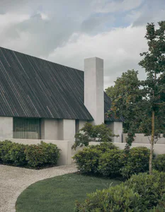 Exterior view of a modern minimalist house featuring light concrete walls, a tall chimney, dark corrugated roof, and a landscaped courtyard garden with gravel paths, manicured shrubs, and a lawn.