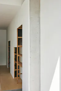 Narrow minimalist hallway featuring built-in oak shelving units recessed into a white wall beside a raw concrete pillar, with light wood flooring.