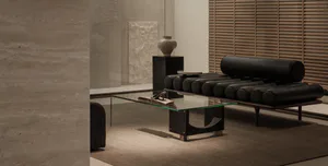Modern minimalist living room featuring a black leather cylindrical bolster daybed, glass coffee table, concrete walls, and a textured stone sculpture on a black pedestal.