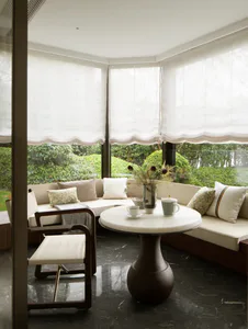 Modern sunroom featuring a curved built-in banquette with neutral cushions, a round white marble table on a dark wood pedestal base, and a wooden armchair with white upholstery, all overlooking a lush green garden through large windows with sheer Roman shades.