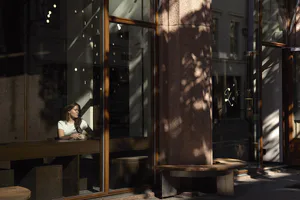 A woman with long brown hair wearing a white t-shirt sits at a wooden counter inside a modern cafe, holding a white coffee cup and looking out the window. Outside, a curved wooden bench sits beside a large textured stone pillar under strong sunlight that casts intricate tree shadows across the glass facade.