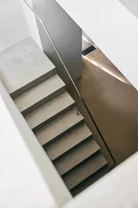 High-angle view of a minimalist concrete staircase with stainless steel handrail and dramatic natural light casting long shadows on the lower landing.