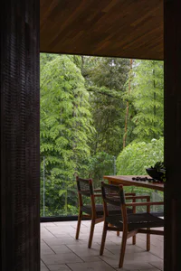 View from inside a wooden structure looking out onto a tiled balcony with teak dining chairs and table, overlooking a dense bamboo and conifer forest.