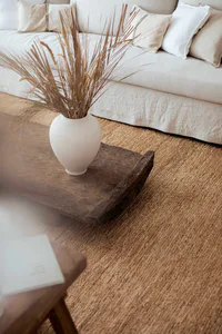 Close-up view of a rustic dark wood live-edge coffee table holding a white ceramic vase with tall dried palm fronds and wheat stalks, set on a natural jute rug in front of a light neutral sofa with linen pillows.