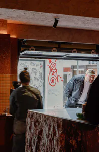 Interior view of a modern cafe showing a textured stone service counter, a barista with hair in a bun facing away, and a bearded man in a cap leaning on the counter. The background features white walls covered in colorful graffiti and red street art, with warm orange lighting and an open pass-through window.