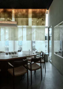 Modern minimalist dining room featuring a long wooden table, woven rattan chairs, large translucent onyx or travertine wall panels, and a striking suspended copper art installation on the ceiling.