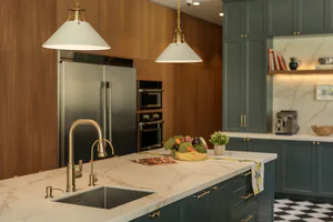 Modern kitchen featuring a large central island with white marble countertop, integrated sink and brass gooseneck faucet, sage green cabinetry, wood panel walls, stainless steel appliances, and black and white checkerboard floor.