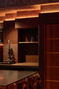 Modern kitchen featuring dark red terrazzo walls and bar counter, wooden cabinetry, brown tile backsplash, industrial-style faucet, and warm ambient lighting from above.