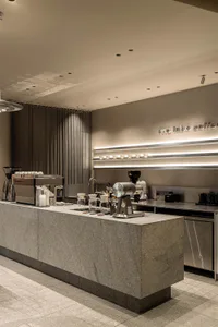 Modern minimalist coffee shop interior with a large grey terrazzo counter, professional espresso machine, coffee grinder, and illuminated shelving displaying white cups.