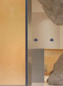 特寫赭黃色肌理灰泥牆，旁邊是藍框玻璃隔板，背景可見抽象混凝土雕塑與幾何藍色牆面細節。