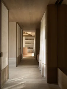 A long, minimalist hallway featuring light wood ceilings and wall paneling, polished concrete floors, sheer white linen curtains, and a view into a study area with built-in shelves.