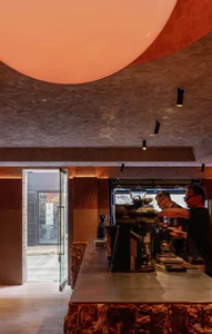 Interior view of a modern cafe featuring a large pink-orange spherical pendant light, textured terrazzo walls and ceiling, a stainless steel coffee bar with professional espresso equipment, and two baristas working. A glass door opens to the street.