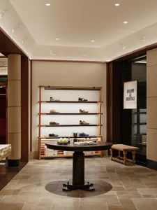 Interior view of a luxury shoe store with multi-tiered wooden display shelves featuring various sneakers, a large dark wood round pedestal table in the center, a wooden bench, stone-tiled floor, and a hanging sign that reads 'BEHAVE AS MORTALS'.