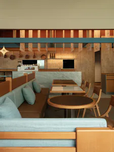 Interior view of a modern cafe featuring a long teal upholstered booth with wooden frame, a round dark wood table, rectangular dining tables with wooden chairs, and a service counter with marble front and hanging copper pans in the background.