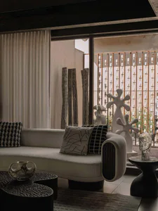 Modern living room featuring a curved bouclé sofa with black and white patterned cushions, sculptural white ceramic artworks, dark textured coffee tables, and a view of an artistic outdoor garden with wooden logs and metal screen.