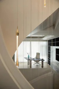 Modern minimalist home office viewed from a curved floating staircase with white marble treads, featuring a black asymmetrical desk, light grey ergonomic office chair, and large sheer-curtained window.