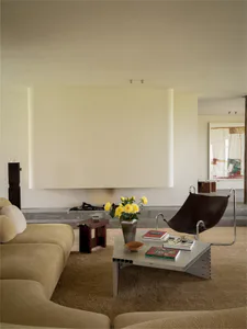 Minimalist living room with beige sectional sofa, brown leather sling chair, metallic coffee table, marble fireplace, and yellow roses in a vase.