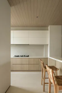 Modern minimalist kitchen with light wood cabinetry, white countertops, a black cooktop, and two wooden bar stools with woven seats in the foreground.