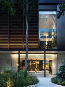 Night view of a modern two-story house with large glass windows, illuminated horizontal wood slats, stone cladding, and a landscaped garden with a tall birch tree.