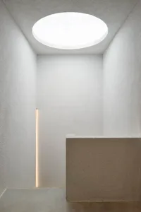 Minimalist concrete stairwell featuring a large circular skylight, vertical LED light strip, and textured plaster walls.