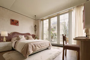 Elegant contemporary bedroom featuring a curved dusty-rose velvet headboard, neutral bedding, textured wallpaper, and large glass doors leading to a balcony with city views.