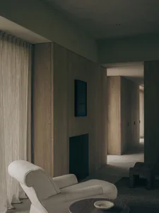 A contemporary minimalist living room featuring light oak wood paneling, a sculptural off-white armchair, a dark wooden side table, and a recessed fireplace niche under soft ambient lighting.