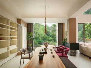 Modern dining room featuring a long live-edge wooden table, pink velvet chairs, built-in shelving, and large windows overlooking a dense green forest.