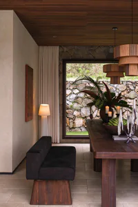Modern rustic dining room featuring a dark wood banquette with upholstered seating, a large wooden dining table, layered wooden pendant lights, a stone wall with large window overlooking a garden, and tropical floral arrangement with candles.