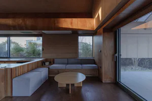 Modern minimalist living room entirely clad in warm plywood with built-in grey upholstered seating, a round wooden coffee table, and large glass doors opening to an outdoor area.