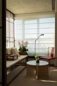 Modern minimalist living room interior with a wooden window seat with cream cushions, a round wooden coffee table, a bright red metal chair, potted orchids, and large windows with sheer blinds overlooking a city skyline.