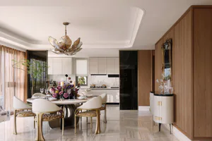 Modern luxury dining room featuring a large round marble dining table, cream upholstered chairs with gold bases, a sculptural metallic flower chandelier, warm walnut wood paneling, and an open view to a white kitchen in the background.