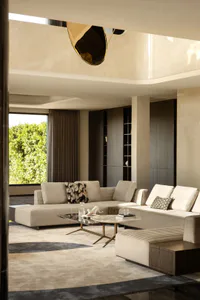 Modern luxury living room with a large beige sectional sofa, marble and glass coffee tables, sculptural brass pendant light, and large window overlooking green foliage.