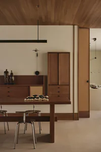 Modern minimalist dining room featuring a solid walnut dining table, matching bench and stools with metal legs, a long black linear pendant light, wooden wall cabinetry, and bronze sculptures on a sideboard against a neutral wall.