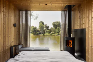 Modern minimalist bedroom with light wood paneling, large picture window overlooking a lake, built-in daybed, and black wood-burning fireplace stove with visible fire.