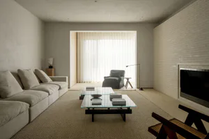 A modern minimalist living room featuring a long beige sectional sofa, a glass and dark wood coffee table on a woven area rug, a white brick fireplace, and a view into a sunlit adjacent space with sheer curtains and a blue armchair.
