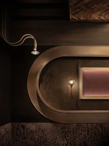 Close-up of a dark curved brass wall panel featuring two spherical wall sconces, a pink velvet artwork, and a textured dark carpet below.