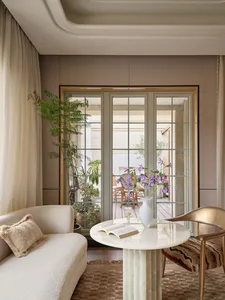 Modern living room interior featuring a curved cream sofa, a round white marble pedestal table with an open book and purple flower vase, a gold metallic chair with striped cushion, and large glass doors opening to a sunlit patio with plants.