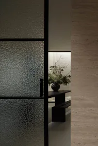 View through a black-framed textured glass door into a minimalist entryway featuring a black console table with a large floral arrangement in a dark vase, set against a textured plaster wall.