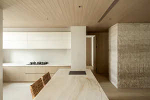 Modern minimalist kitchen with a large travertine stone island, woven leather dining chairs, white handleless cabinetry, gas cooktop, and light wood slat ceiling.