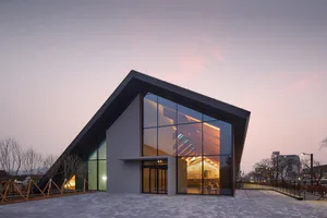 Exterior view of a modern asymmetrical cultural center with large glass walls revealing a warmly lit wooden interior at twilight.