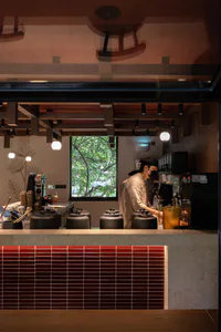 Modern minimalist cafe interior featuring a red tiled counter, warm wooden ceiling beams, spherical pendant lights, and a barista working at the coffee station with a large window showing green trees outside.