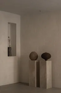 A serene minimalist interior corner with two concrete pedestals holding a textured spherical ceramic vessel and a dark organic-shaped vessel, alongside a recessed niche containing a stone vase with dried botanicals.