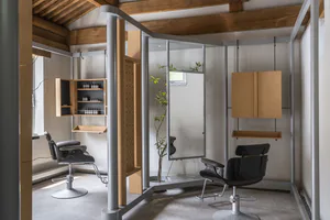 Modern minimalist hair salon interior with two black salon chairs, plywood storage units, steel structural columns, and a large mirror under a pitched wooden ceiling.