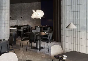Modern restaurant interior with light grey tiled walls, dark round dining tables, grey upholstered chairs, and a large white sculptural pendant lamp hanging over a central table.